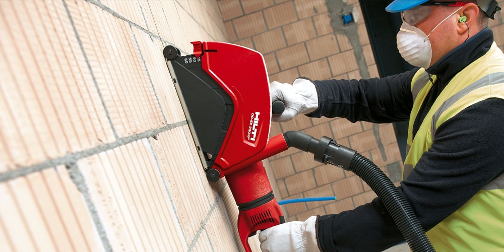 Ein Bild mit einem Arbeiter, der mit einer HILTI Staubhaube (Trennen) DC-EX 230/9" schnei an einer Ziegelwand arbeitet.