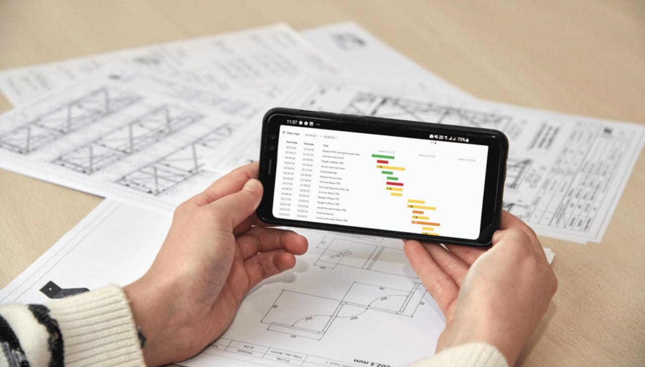Zwei Hände halten ein Smartphone mit Hilti Bausoftware, im Hintergrund liegen Pläne auf einem Tisch.