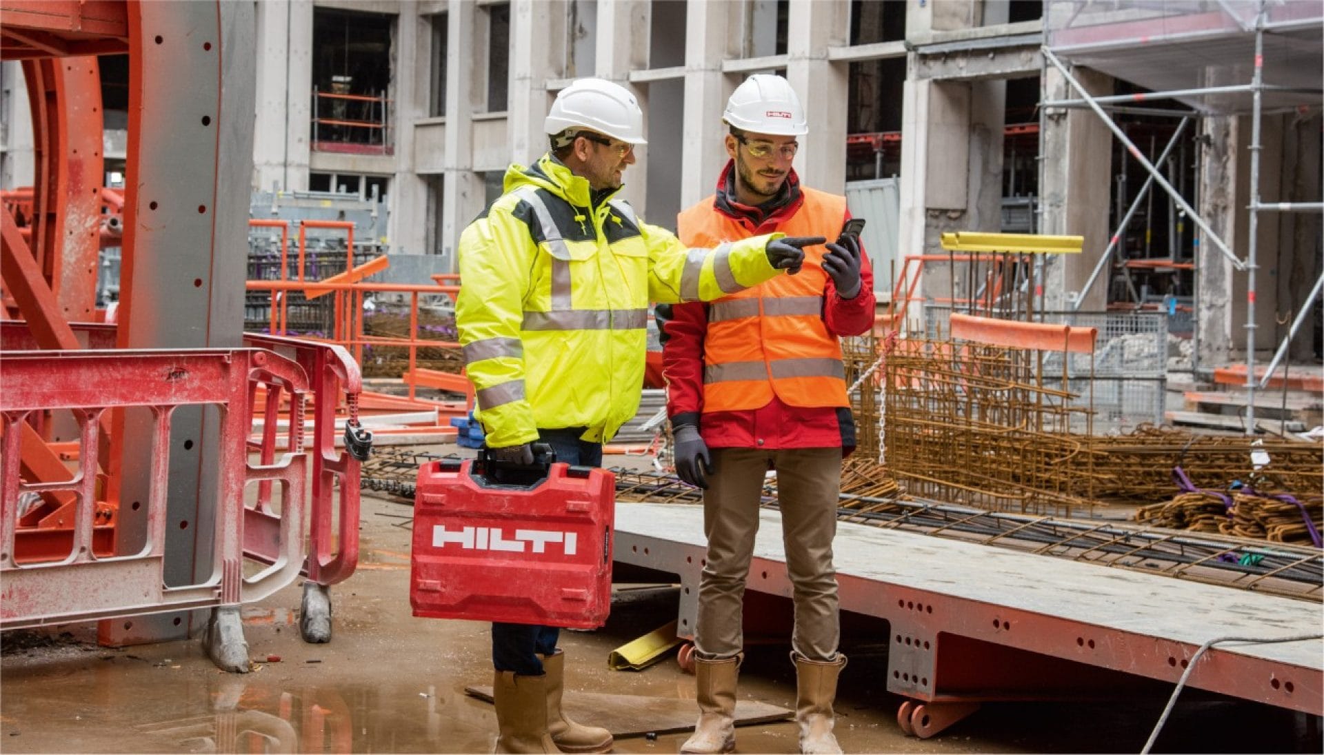 Eine große Baustelle, inmitten der zwei Männer in Warnwesten und Baustellenhelmen stehen. Die beiden sprechen und schauen auf ein Handy, einer davon hält einen roten Hilti Werkzeugkoffer.