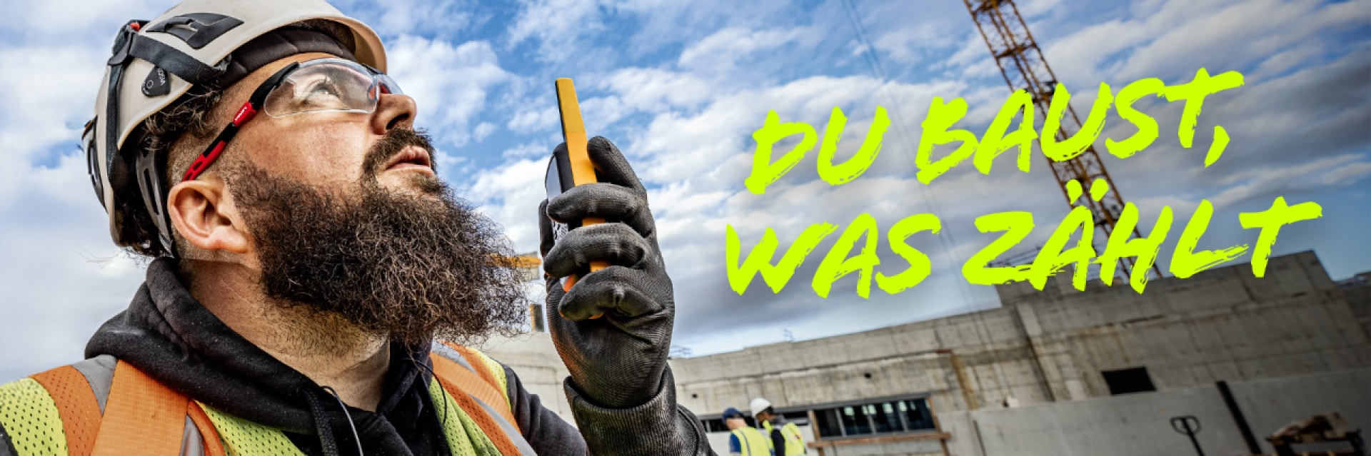 Ein Mann in Schutzkleidung spricht in sein Smartphone, im Hintergrund ist eine große Baustelle.